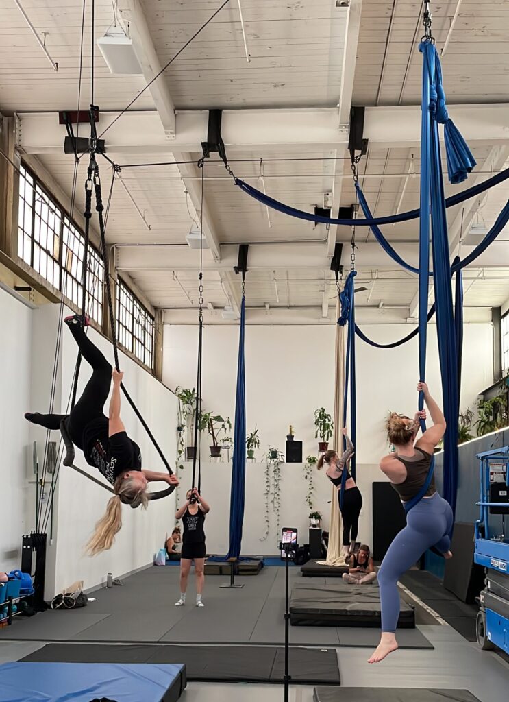 Aerial arts studio showing tall ceiling and multiple apparatuses in us including a long haired person inverted on a trapeze, two people on blue slings, one person on straps, with a couple others in the background.