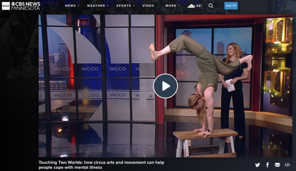 Sherry standing in background as Lynn holds a handstand with staggered legs on her handstand bench at a live interview with WCCO-TV 